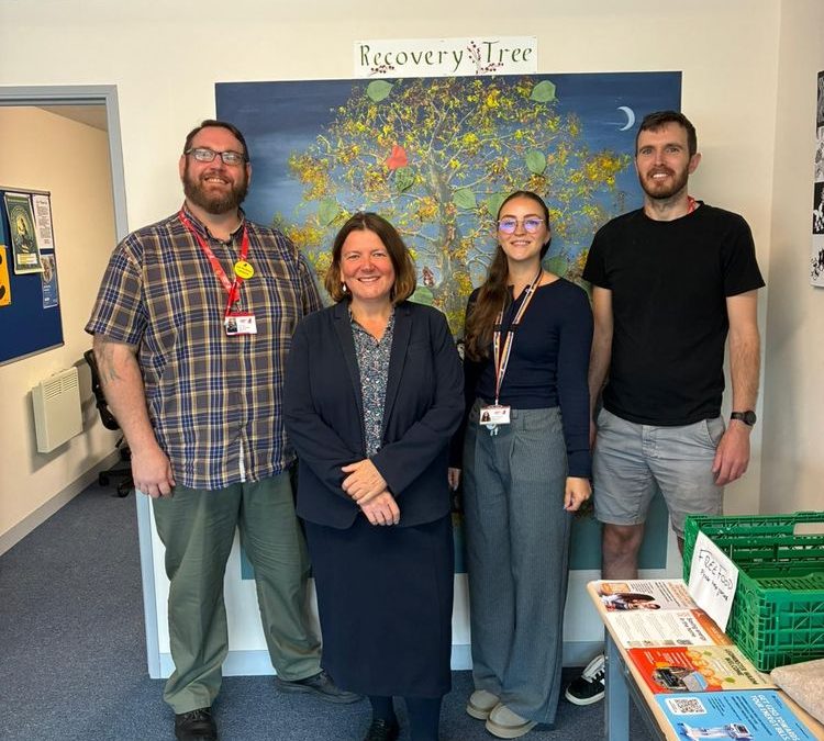Four people including Ellie Chowns stood in front of a painting of a tree titled Recovery Tree