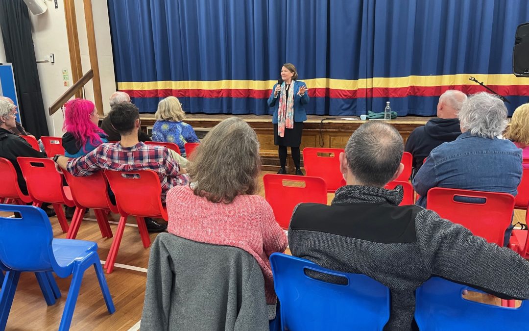 An auditorium with a speakers at the front and an audience sitting on colourful chairs.