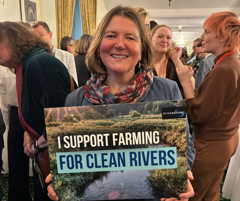MP Ellie Chowns is at the event of River Action, and she holds poster with sentence "I support farming for clean rivers". There are a lot of people behind her, who talking with each other
