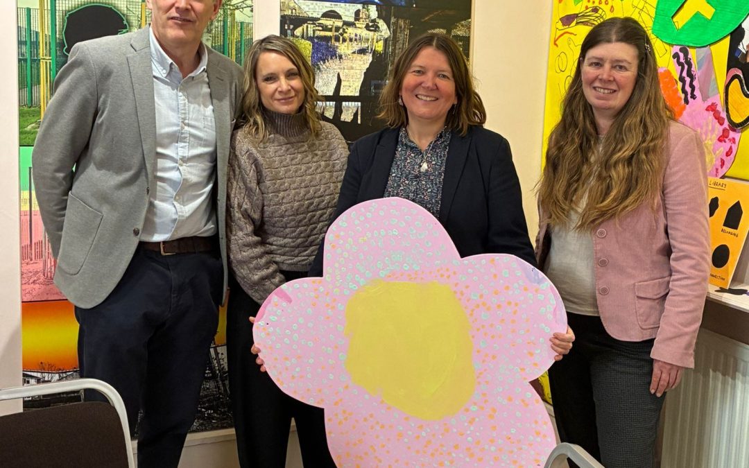 Ellie is standing and holding big paper pink flower. Two ladies and one man is standing next to her. Behind them is crafts form the company she is visiting